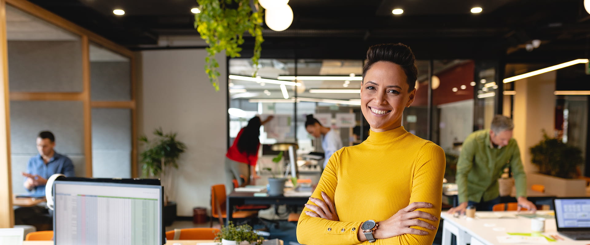 A confident woman in a yellow turtleneck stands with her arms crossed in a vibrant, modern office. 